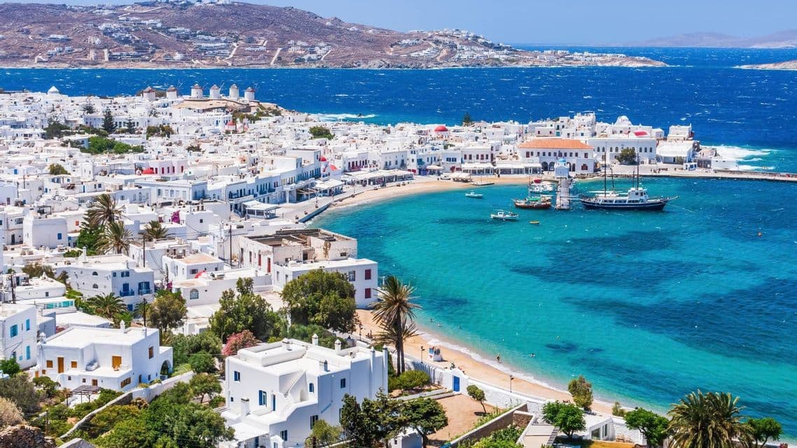 Una vista aerea di una città costiera con edifici bianchi che circondano una piccola spiaggia sabbiosa e una baia turchese con barche.