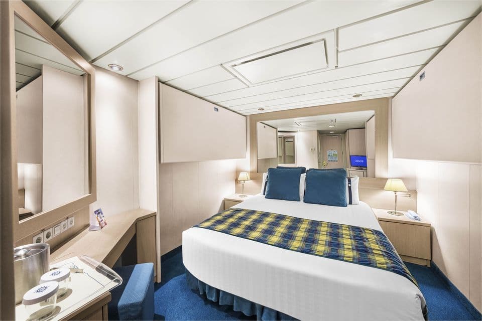 Una cabina compatta su nave da crociera con letto matrimoniale ben fatto, copriletto scozzese, mobile toeletta in legno e moquette blu.