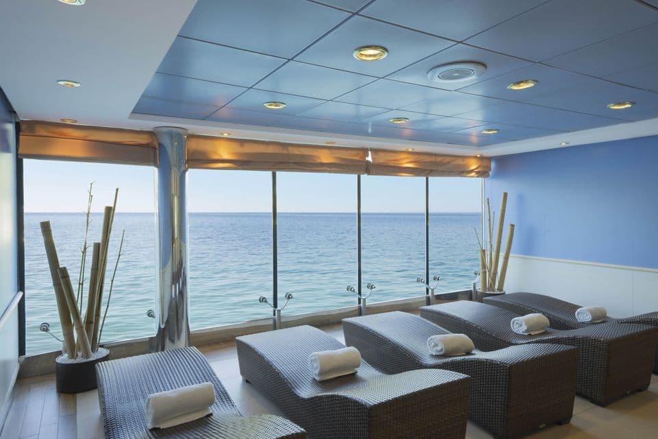 Una fila di lettini intrecciati con asciugamani in una sala relax di una spa si affaccia su un mare blu calmo attraverso ampie finestre.