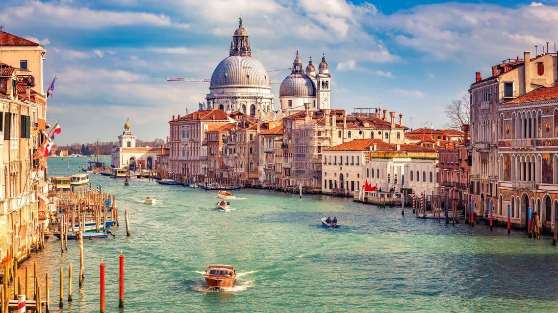 Una veduta diurna di un ampio canale con barche, affiancato da edifici storici e una grande basilica a cupola sullo sfondo.