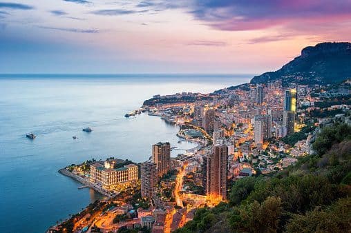 An aerial view of an illuminated coastal city built along the sea at the base of a mountain during sunset.