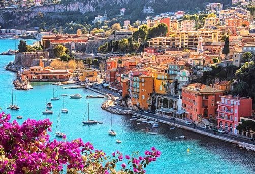 An aerial view of a colorful coastal town with boats docked in a turquoise bay, framed by bright pink flowers.