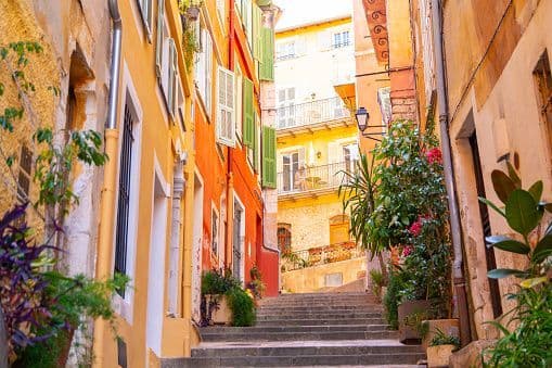 Stone steps lead up a narrow alley flanked by colorful buildings with shutters and potted plants.