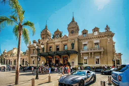 A grand, ornate building with a plaza full of people and luxury cars on a sunny day.