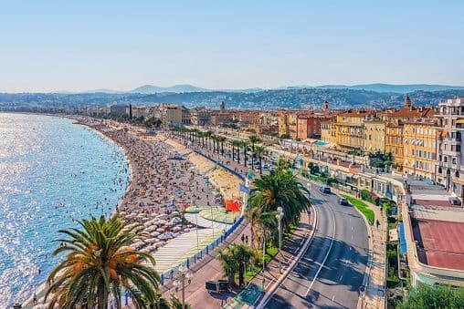 An aerial view of a crowded beach and blue sea along a coastal city with colorful buildings and palm trees.