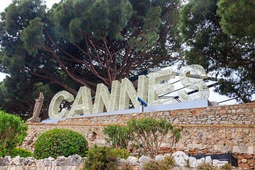 A large sign spelling 'CANNES' in white latticework is displayed on a stone wall in front of a large tree.