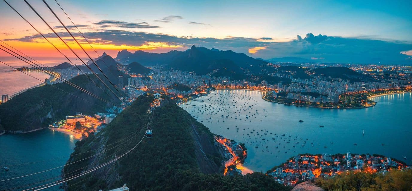 A cable car travels above a mountain, overlooking an illuminated coastal city and a bay filled with boats at sunset.