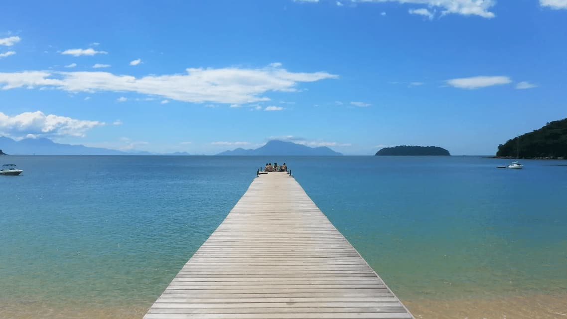 A WeRoad group trip sits at the end of a long wooden pier stretching over a calm blue sea with distant mountains.