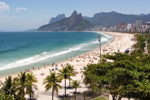 An aerial view of a crowded sandy beach next to a city, with large green mountains in the background under a blue sky.