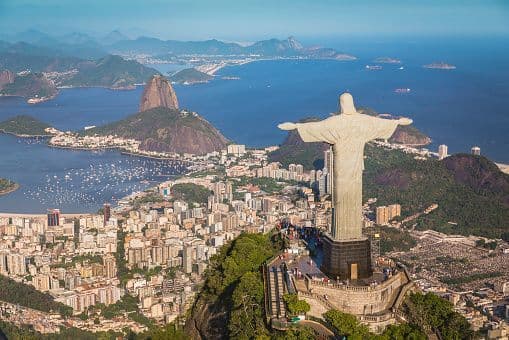 Aerial view of the Christ the Redeemer statue from behind, overlooking a sprawling city, a bay, and Sugarloaf Mountain.