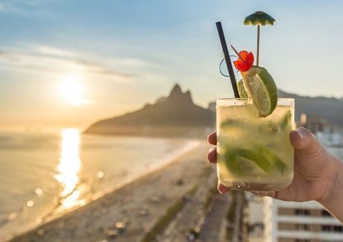 A hand holds a garnished cocktail overlooking a beach with mountains in the background at sunset.