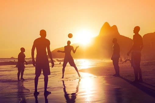 Silhouettes of a WeRoad group trip playing with a ball on the beach at sunset, with mountains in the background.
