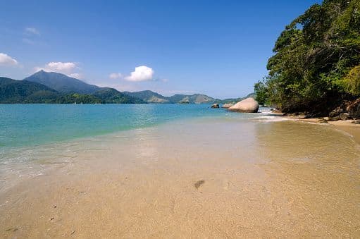 A sandy beach with clear, shallow water curves towards a tree-lined shore, with mountains across the bay under a blue sky.