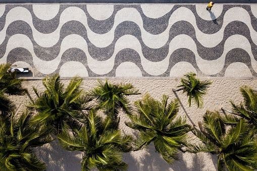 An aerial view of a person walking on a wavy-patterned sidewalk next to a sandy beach with palm trees.