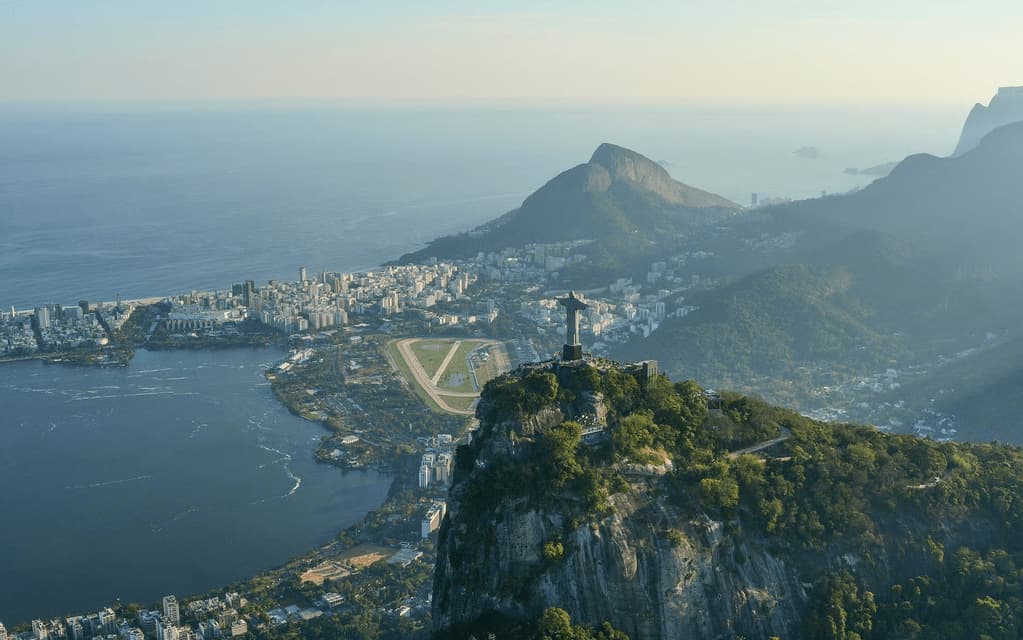 A monumental statue on a high mountain peak overlooks a sprawling coastal city with a lagoon and the ocean.