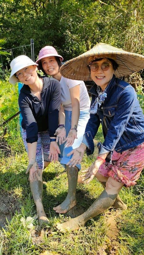 Un voyage de groupe WeRoad de trois femmes souriantes, montrant leurs mains et jambes couvertes de boue dans un champ herbeux.