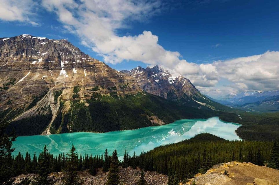 Un vivace lago turchese si snoda attraverso una valle di montagna ricca di foreste di pini, sotto un cielo azzurro con nuvole bianche.