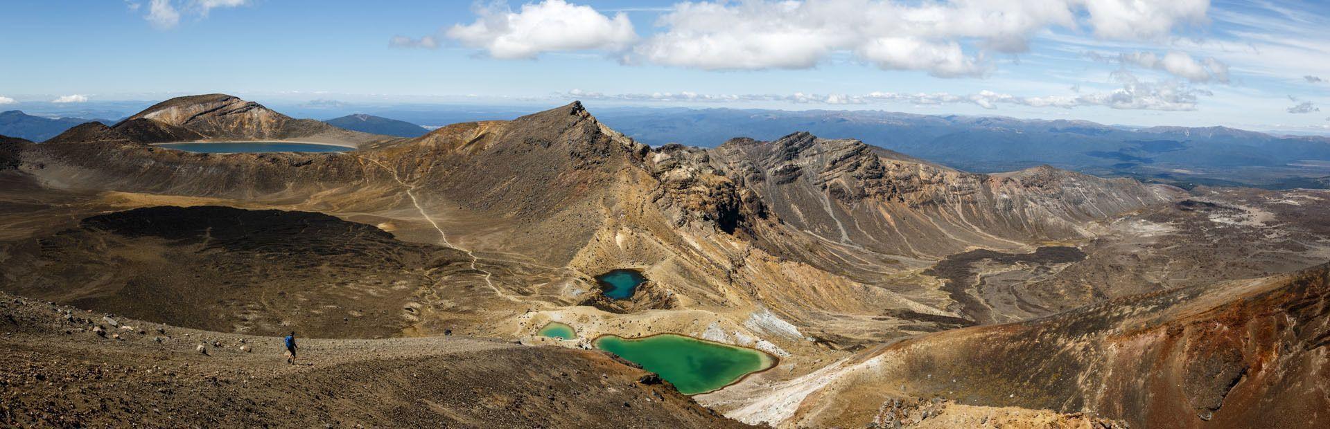 A panoramic view of a volcanic landscape with several green and blue crater lakes, with a hiker walking on a trail in the foreground.