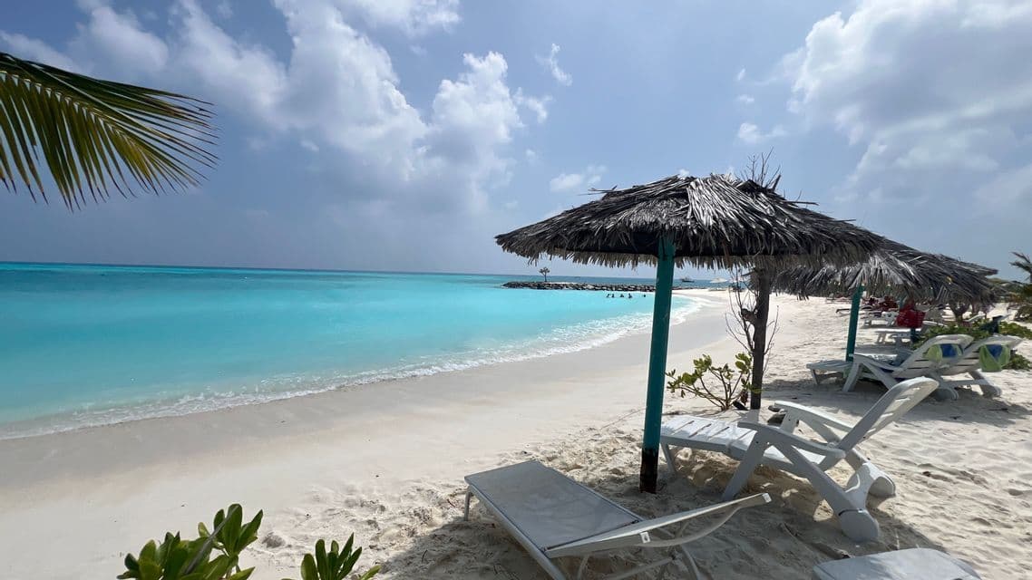 Liegestühle und Strohschirme an einem weißen Sandstrand blicken auf ruhiges, türkisfarbenes Wasser unter einem teilweise bewölkten Himmel.