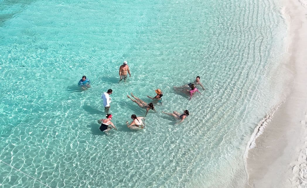 Eine Luftaufnahme einer WeRoad Gruppenreise, die in klarem, flachem türkisfarbenem Wasser an einem weißen Sandstrand schwimmt und entspannt.