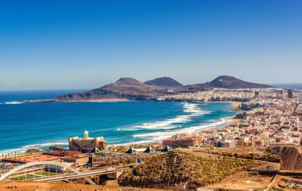 Una veduta dall'alto di una città costiera con una lunga spiaggia sabbiosa, un mare azzurro intenso e montagne aride sotto un cielo limpido.