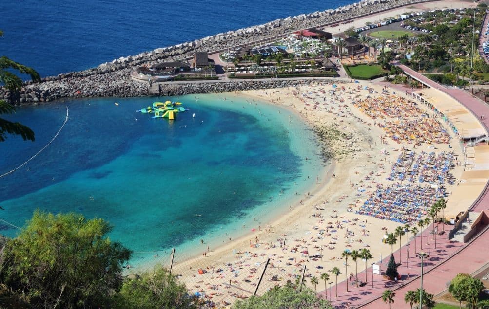 Vista aerea di una spiaggia affollata a forma di mezzaluna, dove le persone prendono il sole sulla sabbia e nuotano nelle acque calme e turchesi.