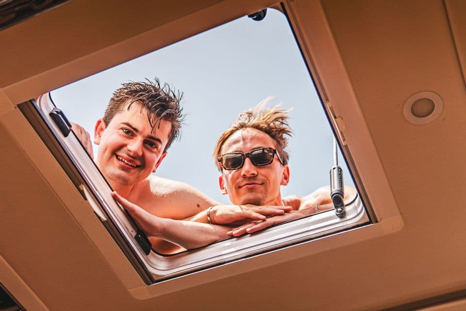 Zwei Männer auf einer WeRoad Gruppenreise, von unten gesehen, lächeln, während sie durch eine offene Dachluke blicken.