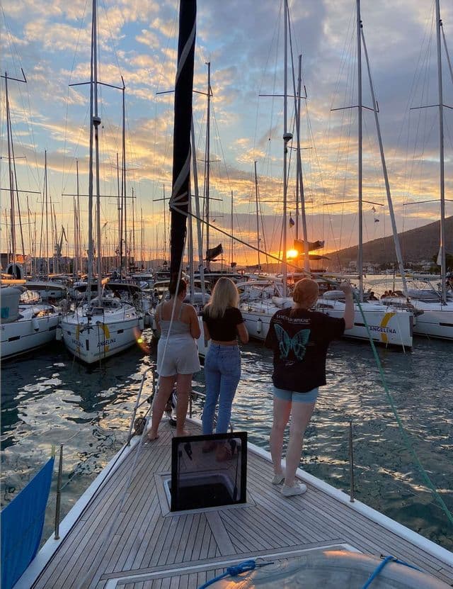 Drei Frauen einer WeRoad Gruppenreise stehen bei Sonnenuntergang am Bug eines Segelbootes in einem mit Booten gefüllten Yachthafen.