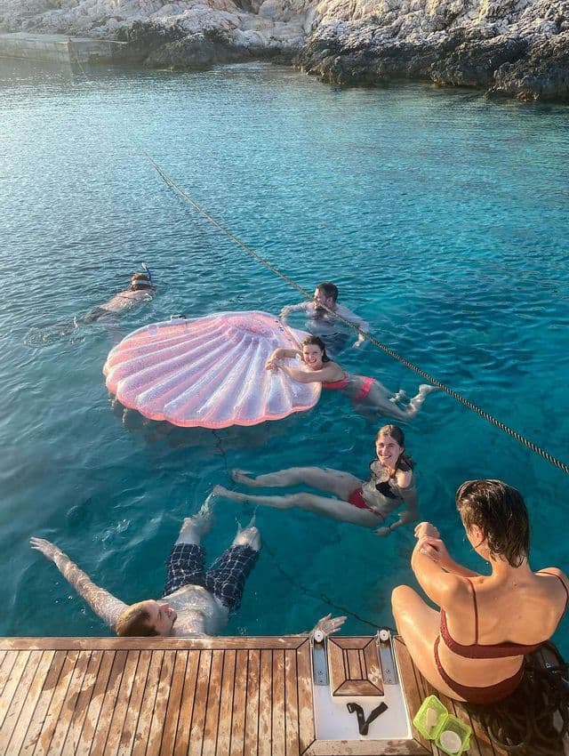 Eine WeRoad-Gruppe badet in klarem, türkisfarbenem Wasser und entspannt auf einem rosafarbenen, muschelförmigen Schwimmtier neben einem hölzernen Bootsdeck.