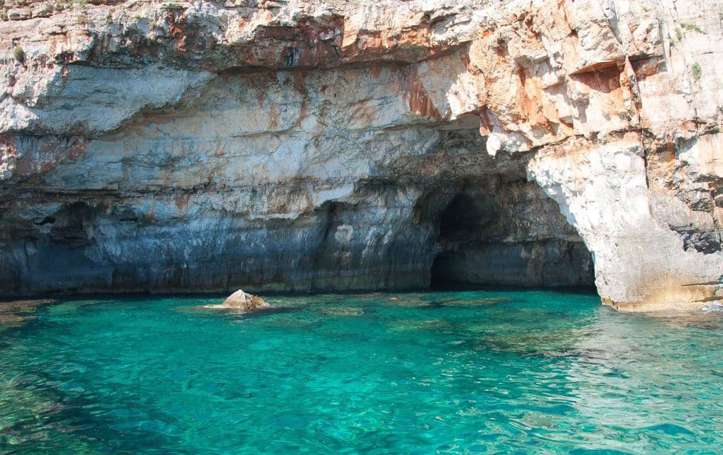 A sea cave at the base of a rocky cliff with clear, turquoise water in the foreground.