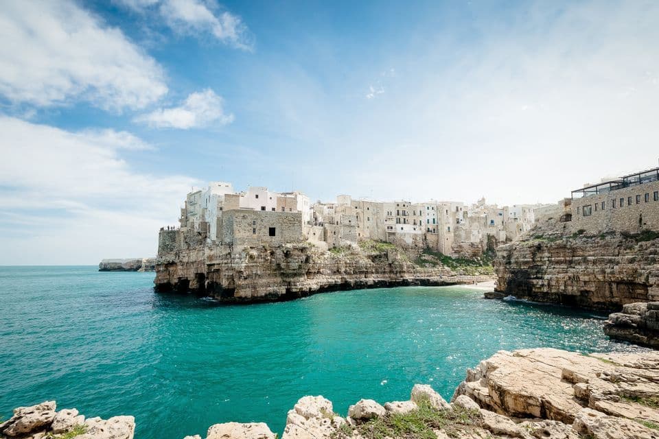 A town with white and stone buildings sits atop a rocky cliff overlooking a turquoise bay under a partly cloudy blue sky.