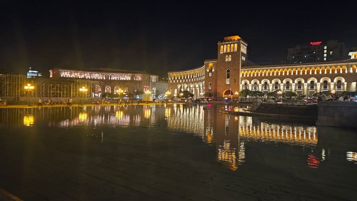 Nachts beleuchtete, verzierte Gebäude mit Bogenfenstern, deren goldene Lichter sich in einem großen Wasserbecken auf einem Stadtplatz spiegeln.