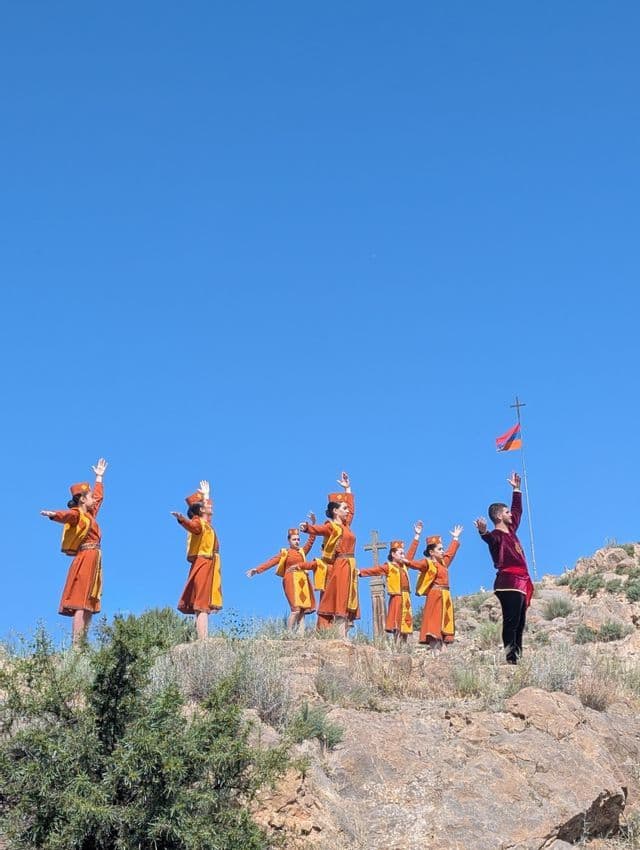 Eine Gruppe Tänzer in traditionellen orangefarbenen Kostümen tanzt auf einem felsigen Hang mit einer armenischen Flagge vor einem klaren blauen Himmel.