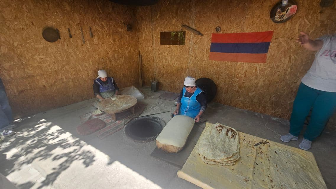 Zwei Frauen bereiten traditionelles Lavash-Brot in einem Raum mit einem Lehmofen zu, während eine armenische Flagge an der Holzwand hängt.