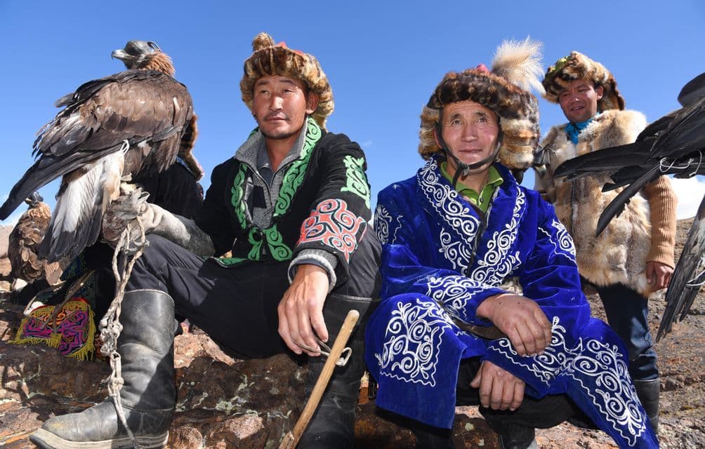 Trois chasseurs d'aigles, vêtus de chapeaux de fourrure traditionnels et de vêtements brodés, sont assis sur des rochers avec leurs aigles royaux sous un ciel clair.