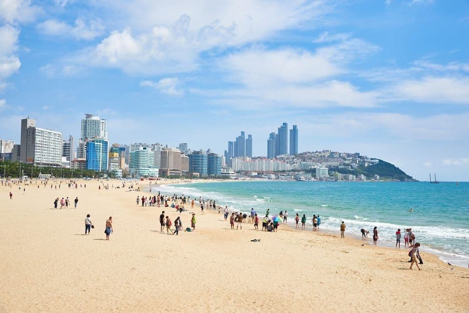 Spiaggia sabbiosa affollata con lo skyline di una città moderna oltre l'acqua sotto un cielo azzurro.