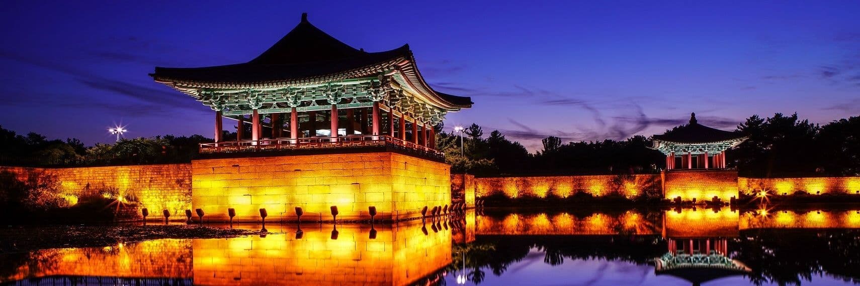 Una tradizionale pagoda asiatica e un muro di pietra illuminati al tramonto, con le loro luci calde che si riflettono nell'acqua immobile.