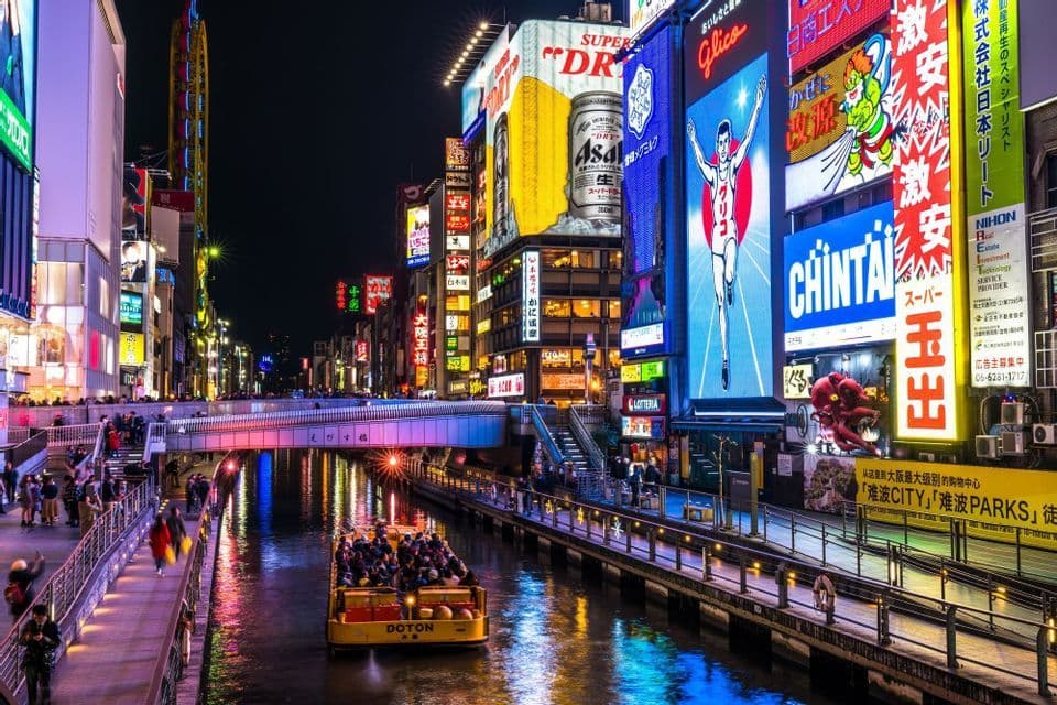 Una barca turistica piena di persone naviga lungo un canale cittadino di notte, circondata da edifici con grandi e vivaci insegne pubblicitarie al neon.