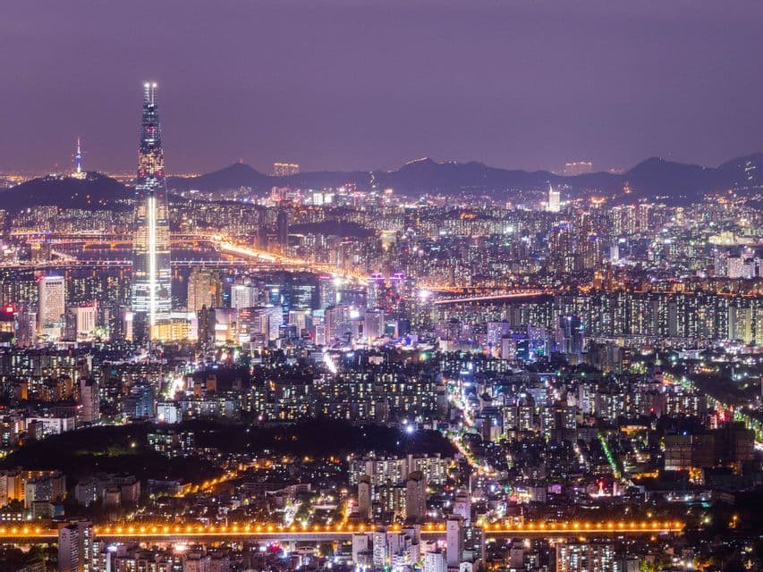Vista aerea dello skyline tentacolare di una città di notte, illuminata da milioni di luci, con un grattacielo imponente e montagne in lontananza.