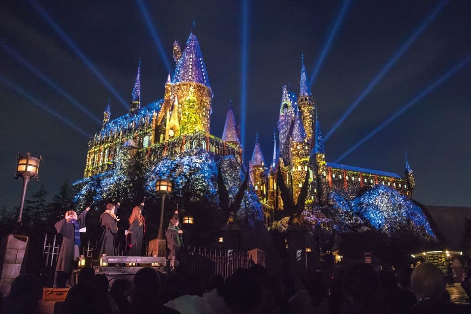 Un gruppo di persone in tuniche su un palco punta bacchette verso un grande castello illuminato di notte da uno spettacolo di luci colorate.