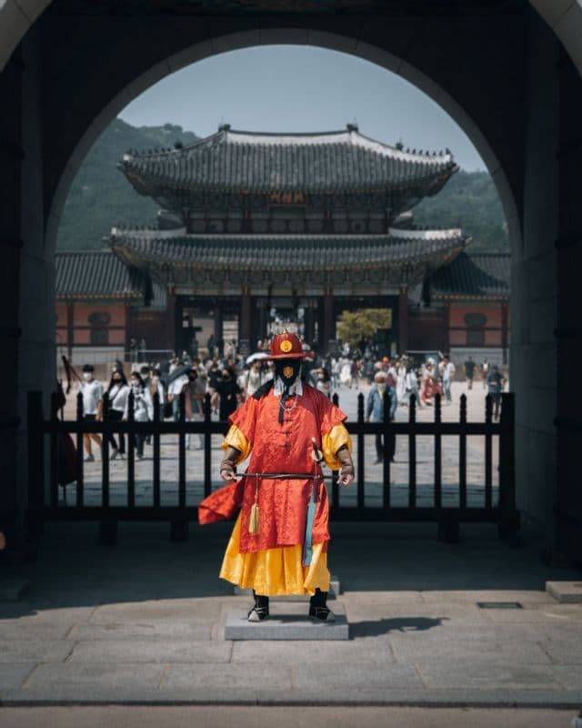 Una guardia in abiti tradizionali coreani rossi e gialli veglia sotto un arco in pietra, con un portone del palazzo sullo sfondo.
