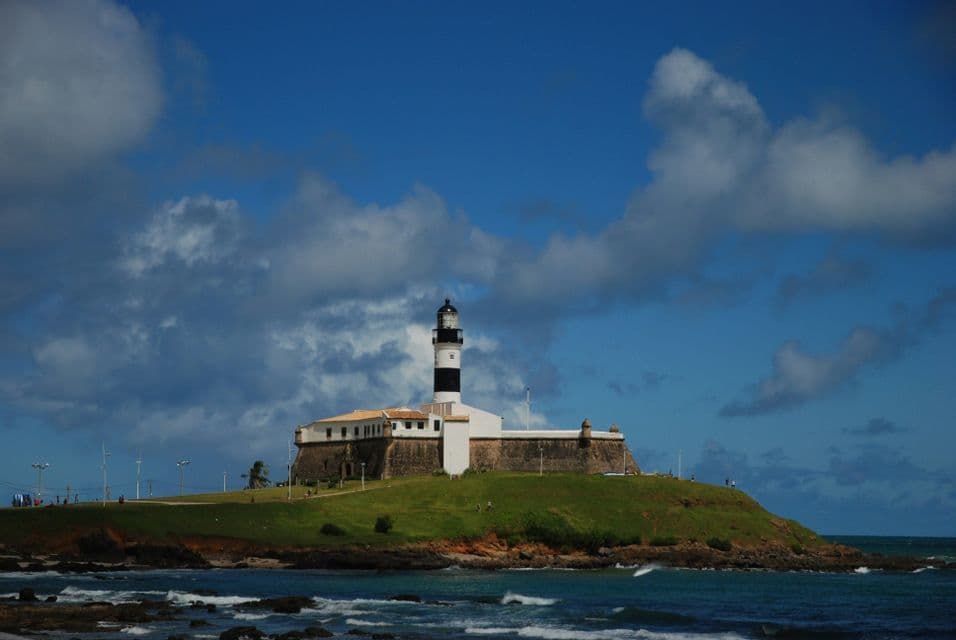 Un forte storico con un faro a strisce bianche e nere si erge su una collina erbosa affacciata sull'oceano in una giornata parzialmente nuvolosa.