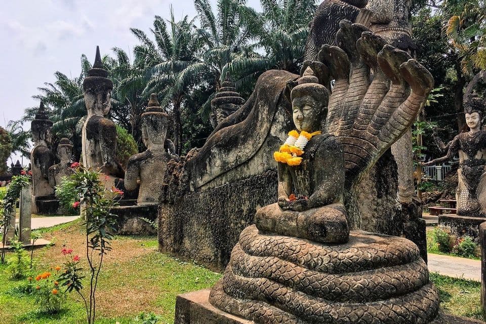 Una statua in pietra del Buddha, adornata con una ghirlanda di fiori, è posta su una base a forma di naga attorcigliato, all'interno di un parco di sculture all'aperto tra le palme.