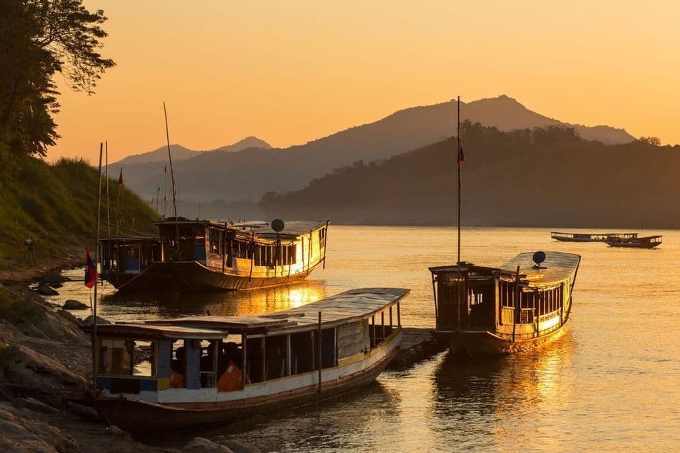 Barche di legno sono ormeggiate lungo un fiume al tramonto dorato, con le montagne che si stagliano sullo sfondo.