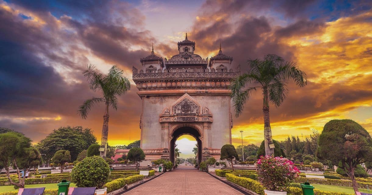 Un grande e ornato monumento ad arco si erge in un parco pubblico, circondato da palme, sotto un drammatico cielo al tramonto.