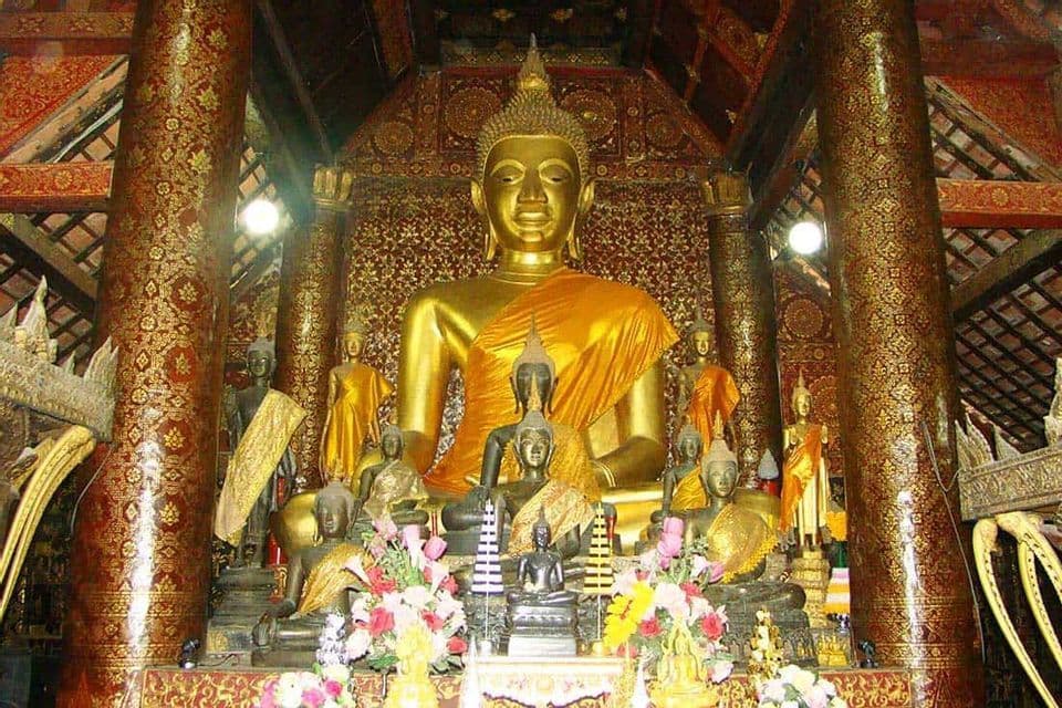 Una grande statua dorata del Buddha è collocata tra figure minori e offerte floreali, all'interno di un tempio sontuoso con colonne rosse e dorate.