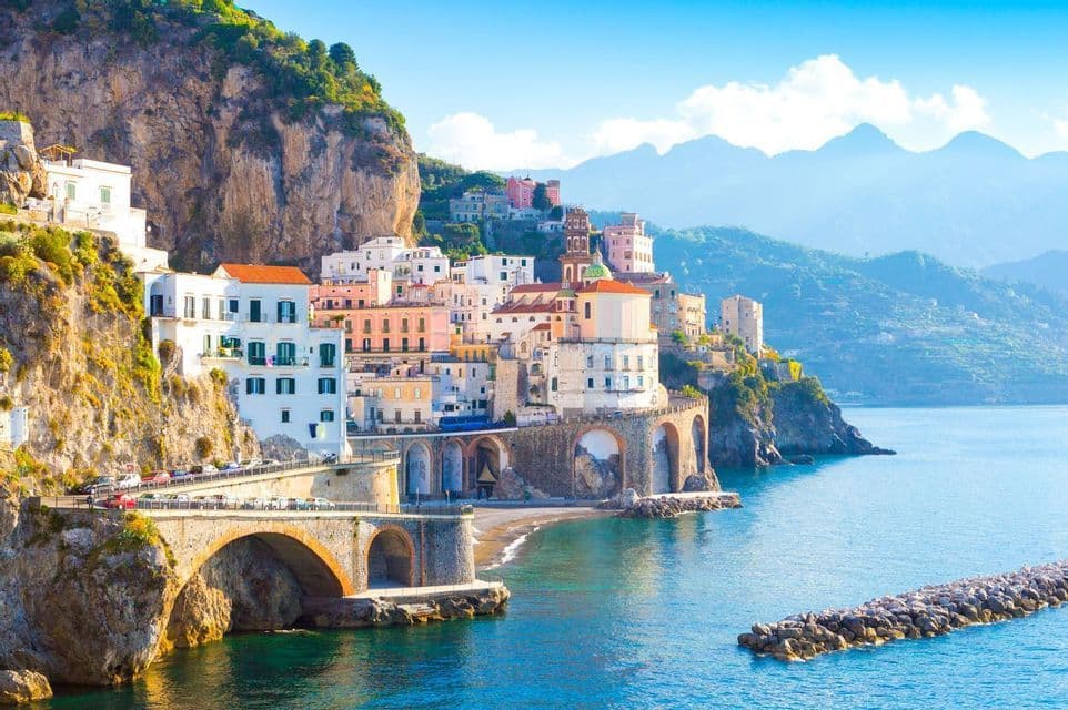 Un pueblo costero con edificios coloridos construidos en un acantilado escarpado, con un puente de arco de piedra sobre el mar azul.