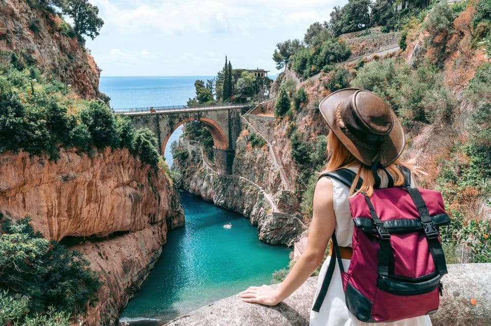 A woman with a hat and backpack looks out at a deep gorge with turquoise water, a stone bridge, and the sea in the background.