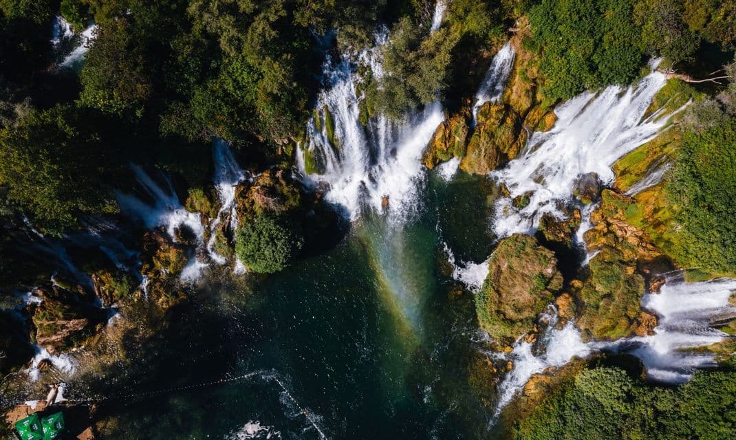 Vista aerea di cascate che si gettano su rocce e vegetazione in una profonda pozza turchese, con un arcobaleno visibile nella nebbia.