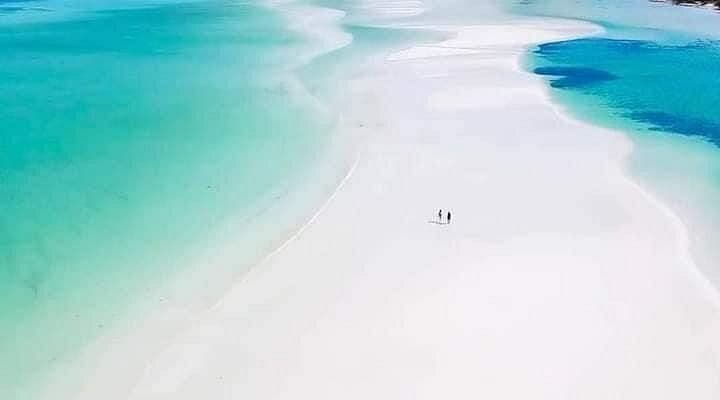 Veduta aerea di due persone che passeggiano su un vasto banco di sabbia bianca circondato da acque turchesi poco profonde.
